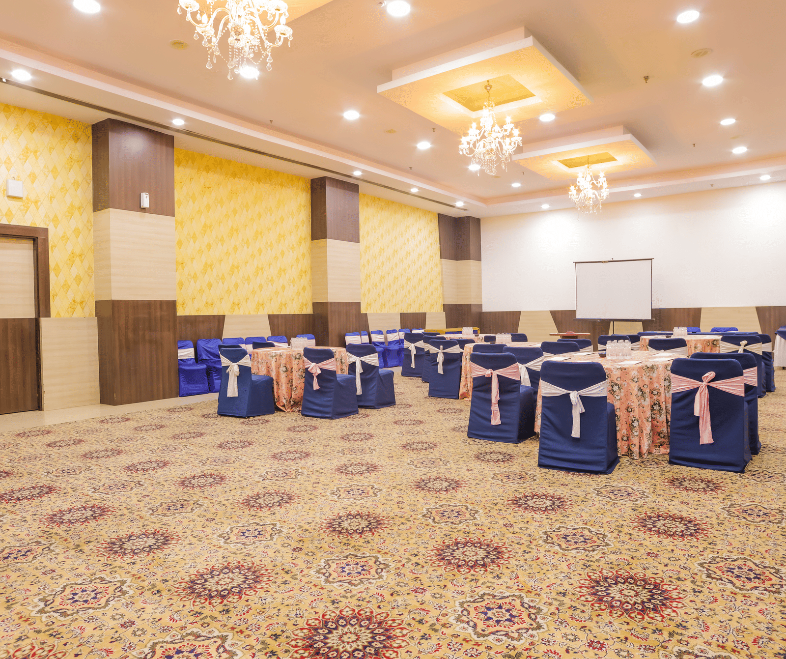 A spacious conference or event room with a patterned carpet, featuring chairs draped in blue covers arranged around tables, and a stage area.