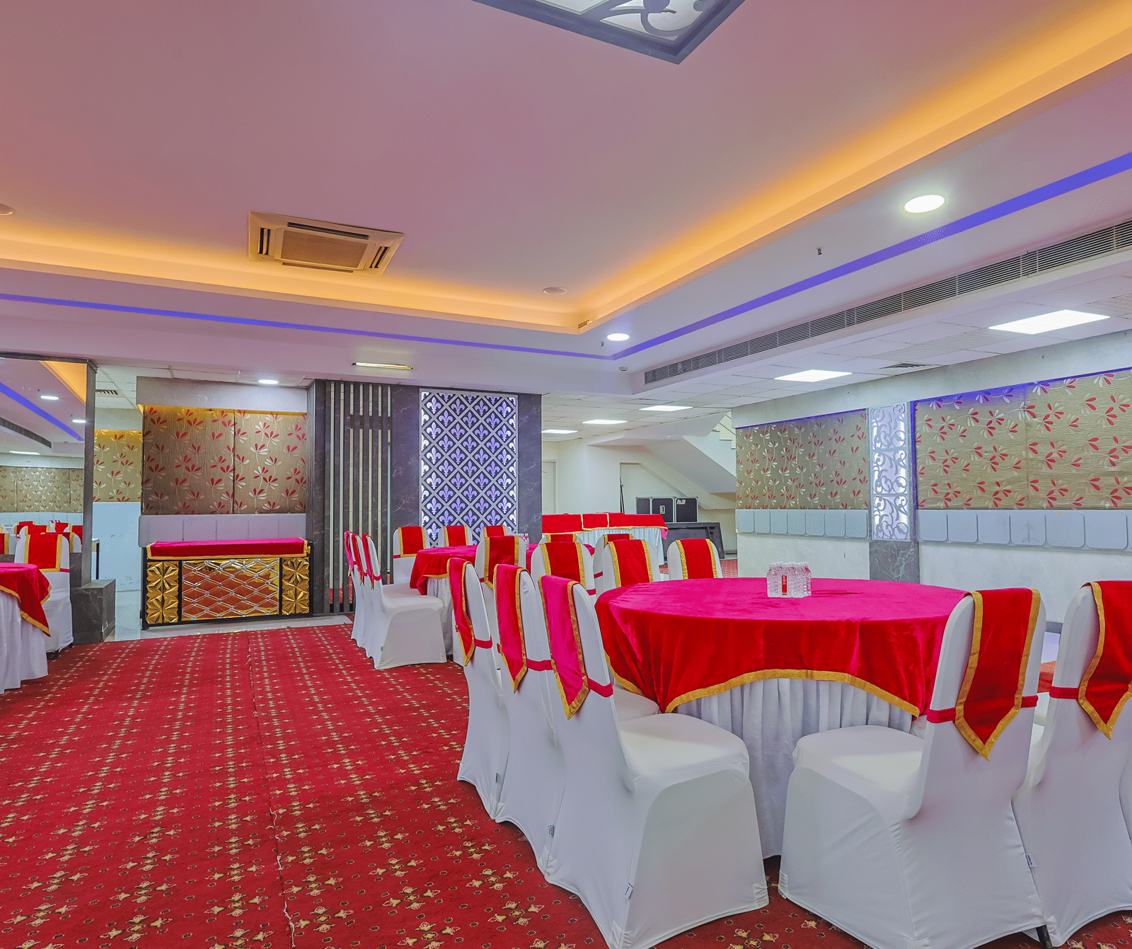  banquet hall set up for an event, featuring tables with white chairs and red tablecloths, arranged on a red patterned carpet.