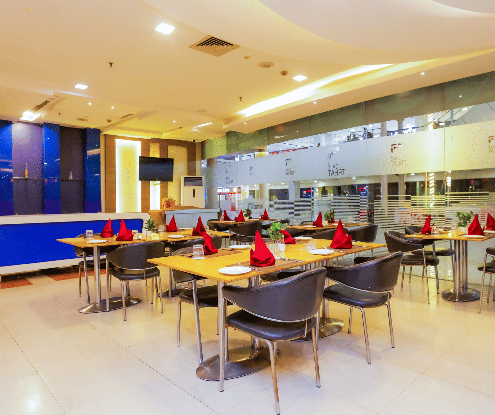 A modern restaurant or lounge area with multiple tables and chairs, a counter or bar area in the background, and bright overhead lighting.