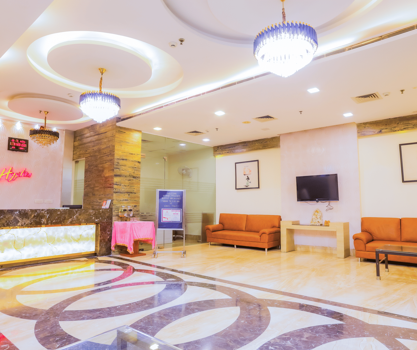 Hotel lobby area with a reception desk, sofas, and wall-mounted TV at Hyphen Premier Hotel, Meerut.