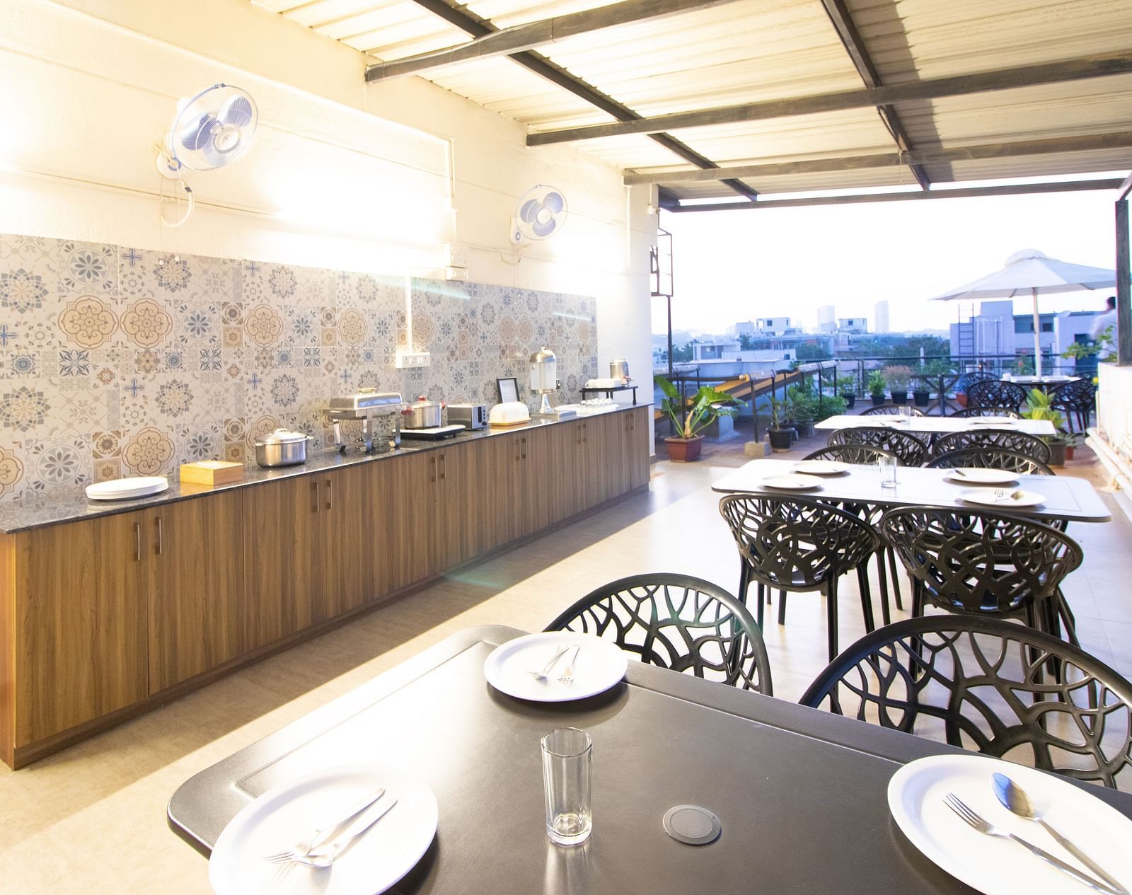 A dining space with tables and chairs under a partial roof, featuring a buffet setup along one side at Sanctum Suites Indiranagar, Bangalore.