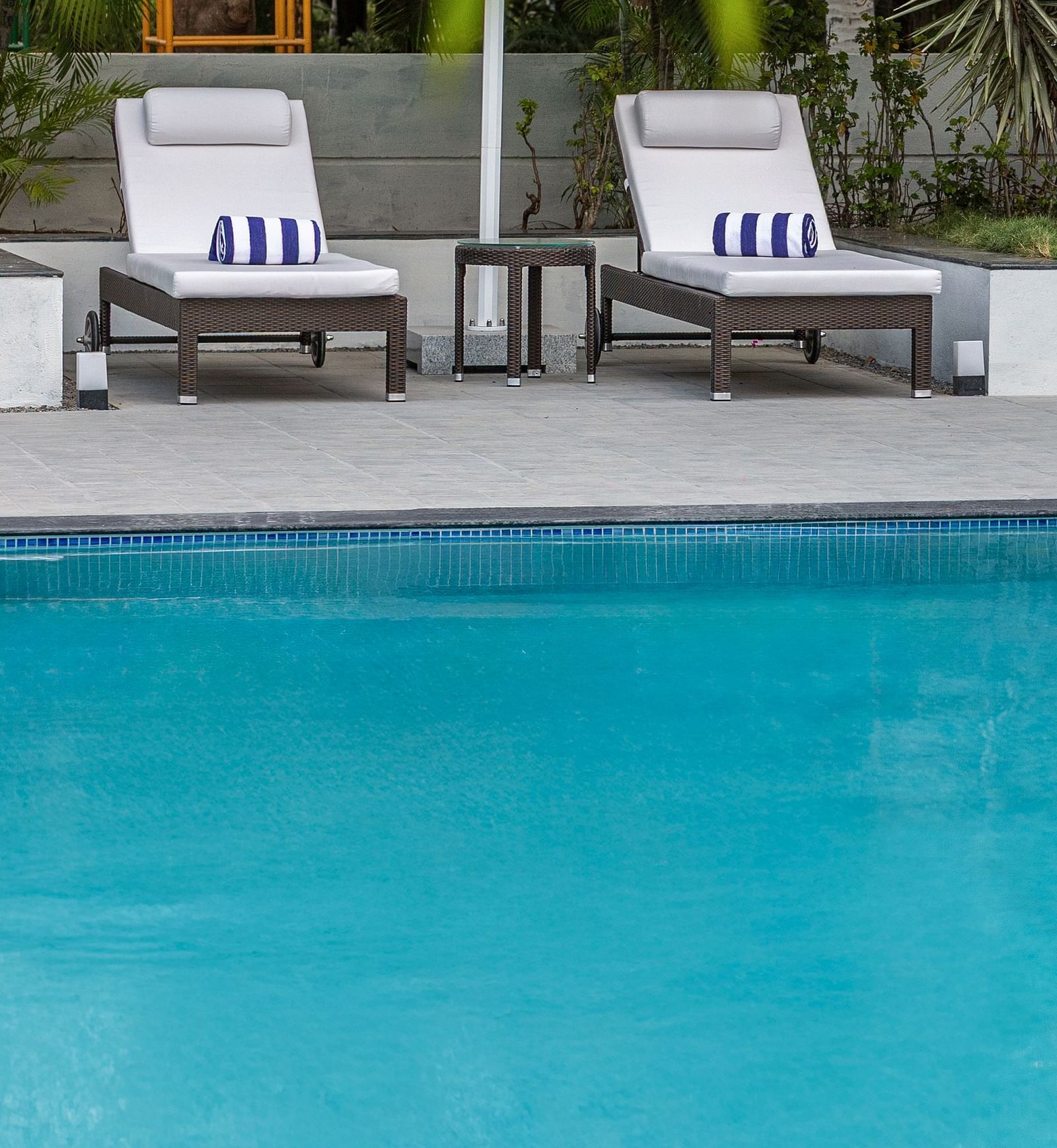Two lounge chairs with striped cushions and a white umbrella by a bright blue swimming pool, surrounded by palm trees - Mango Hill Resort, Coimbatore