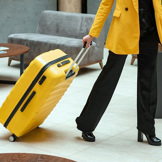 A person in a yellow jacket and black trousers pulling a bright yellow suitcase through a modern hotel lobby.