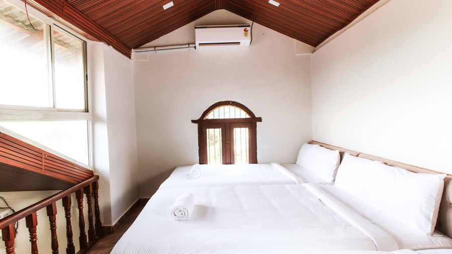A loft style bedroom under a sloped wooden ceiling with white bedding and a wooden staircase railing | Vintage Trails By Ilara
