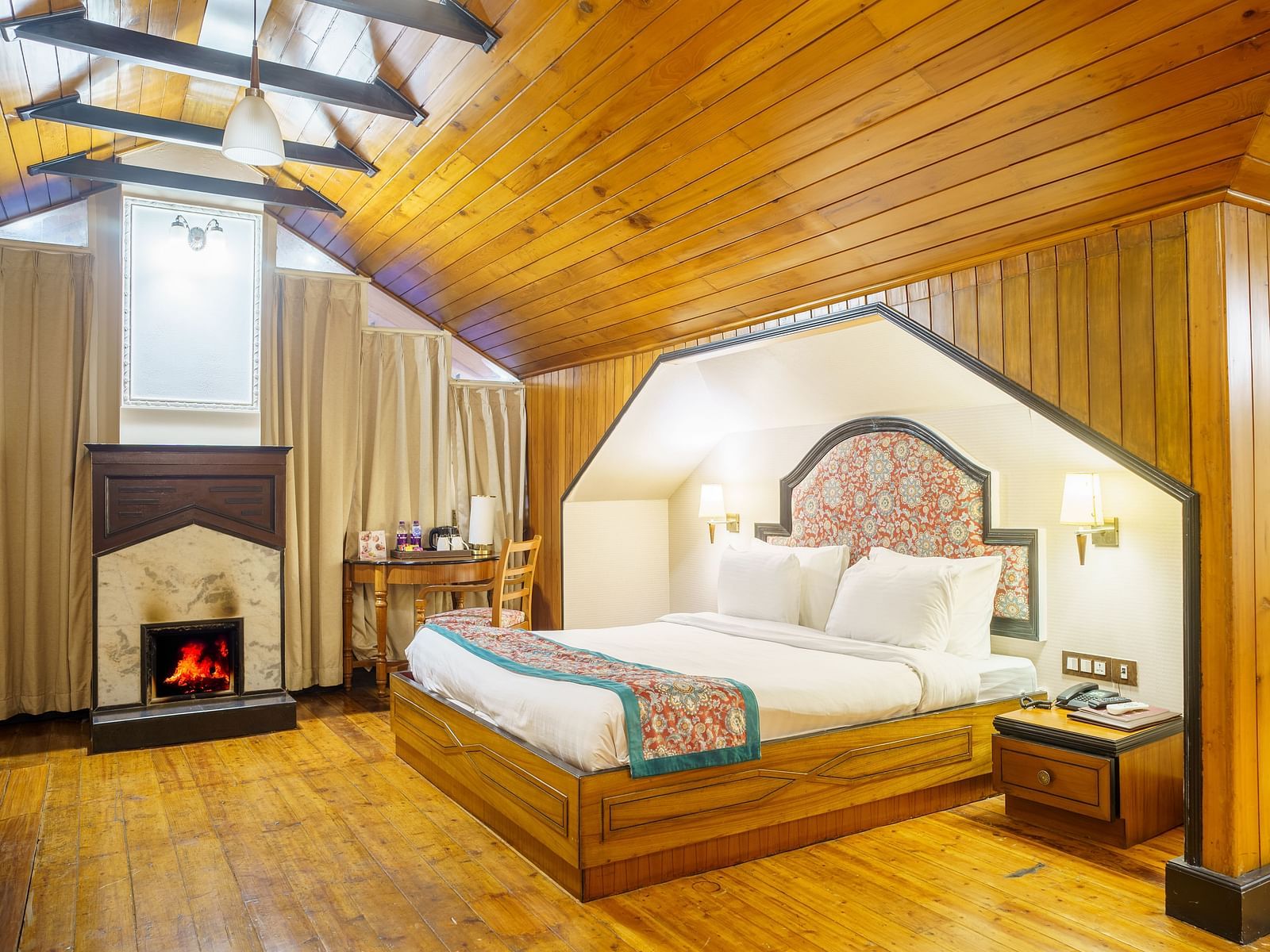 A room at Orsino Spa Resort with a pitched wooden ceiling, a bed with a patterned throw, and a small fireplace.