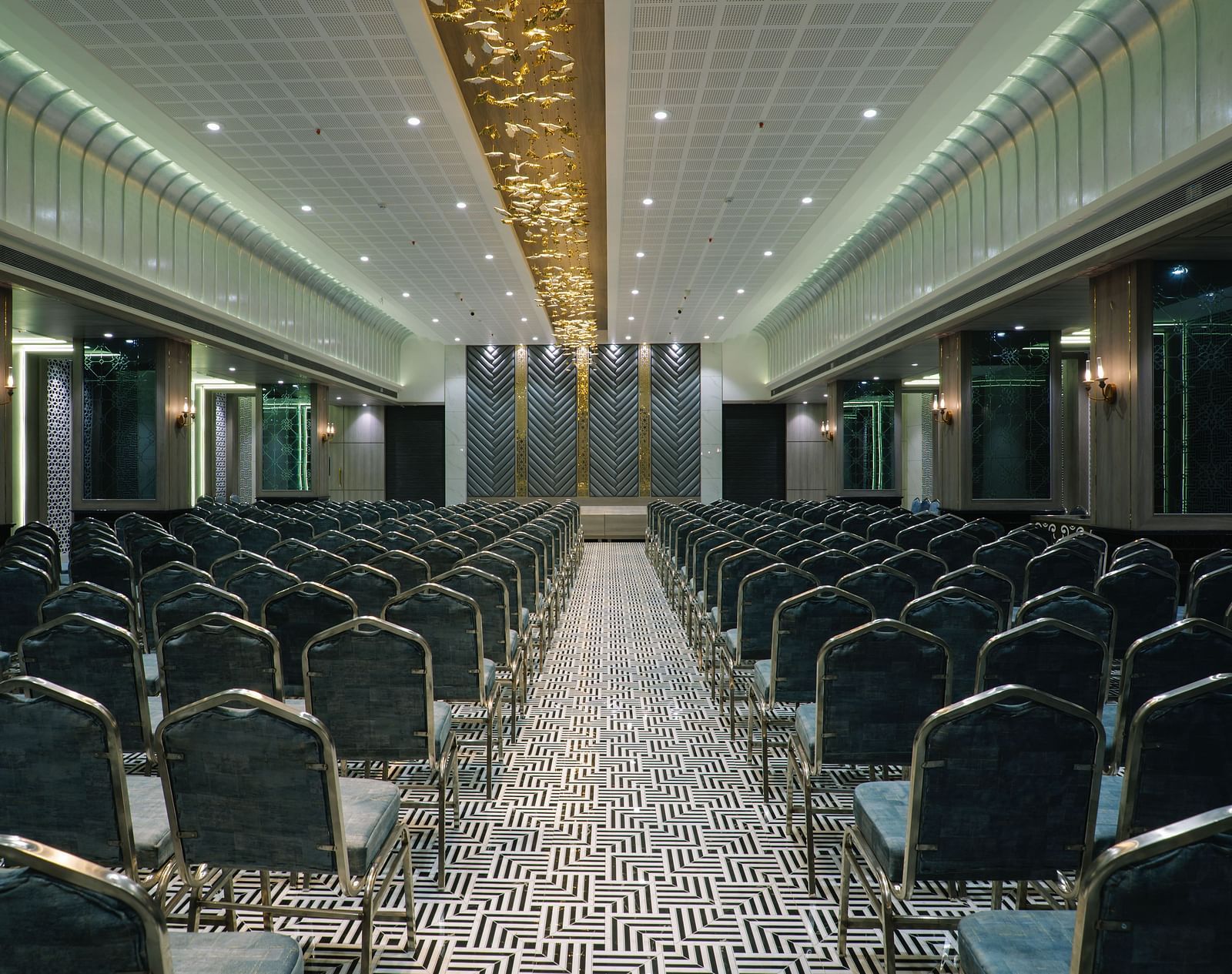 A large room is set up with rows of dark chairs facing a stage or presentation area - Melior Inn