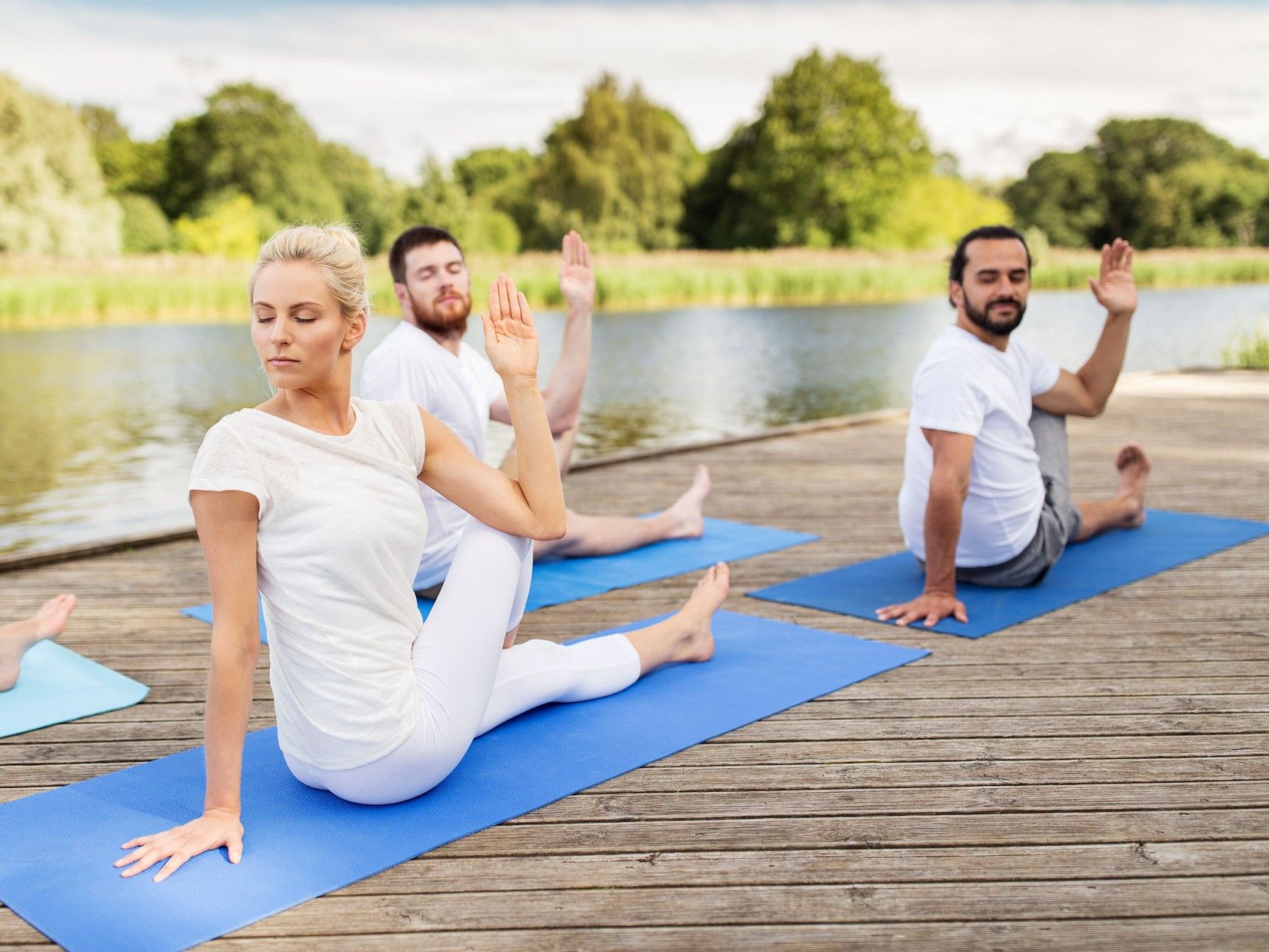 Group of people practicing seated yoga twists on blue mats on a wooden deck by a lake, surrounded by lush greenery at YO1 Longevity and Health Resorts, Catskills.