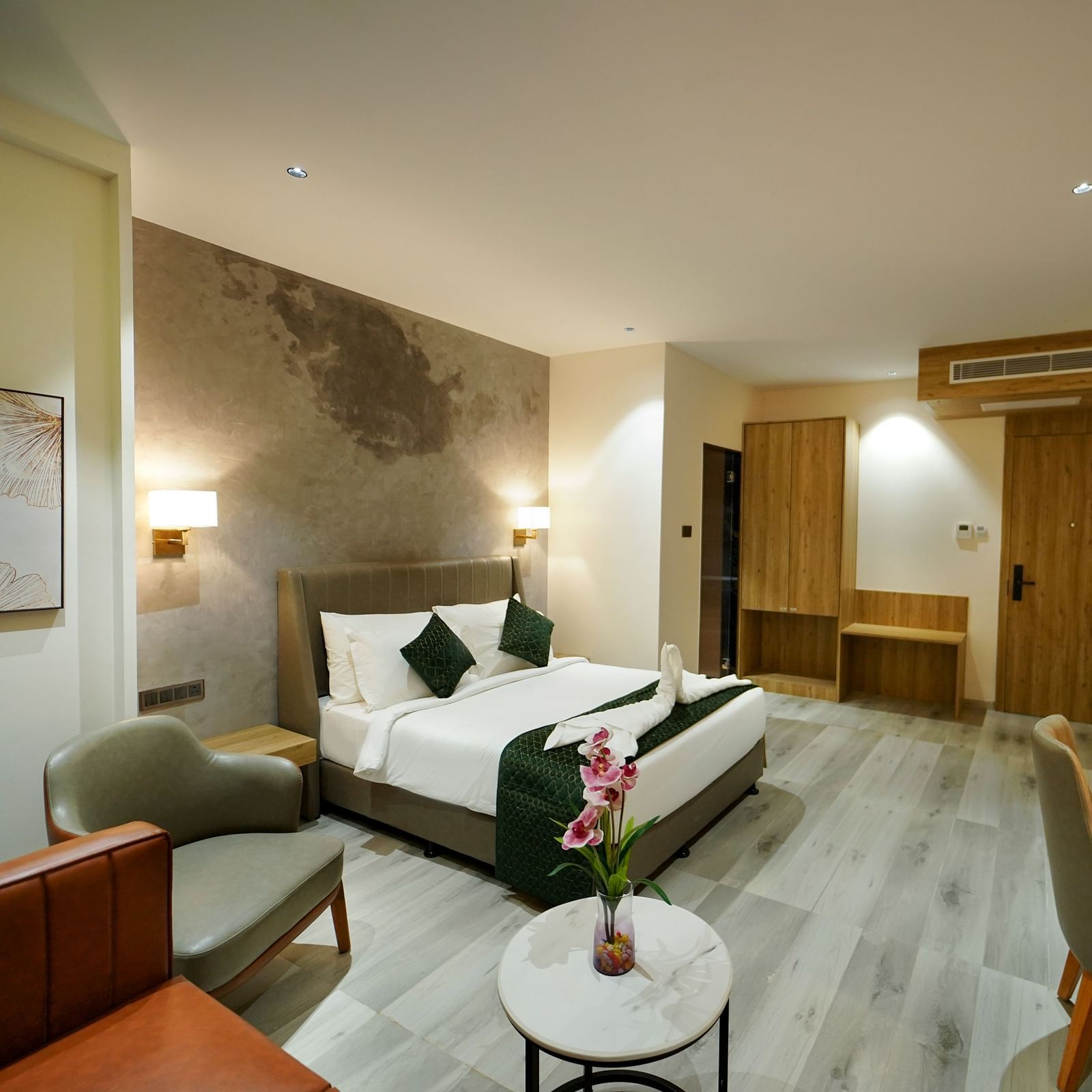 A spacious hotel room includes a bed with pillows, a seating area with chairs and a small table with flowers, and a desk area - Hotel Park Inn, Nellore.