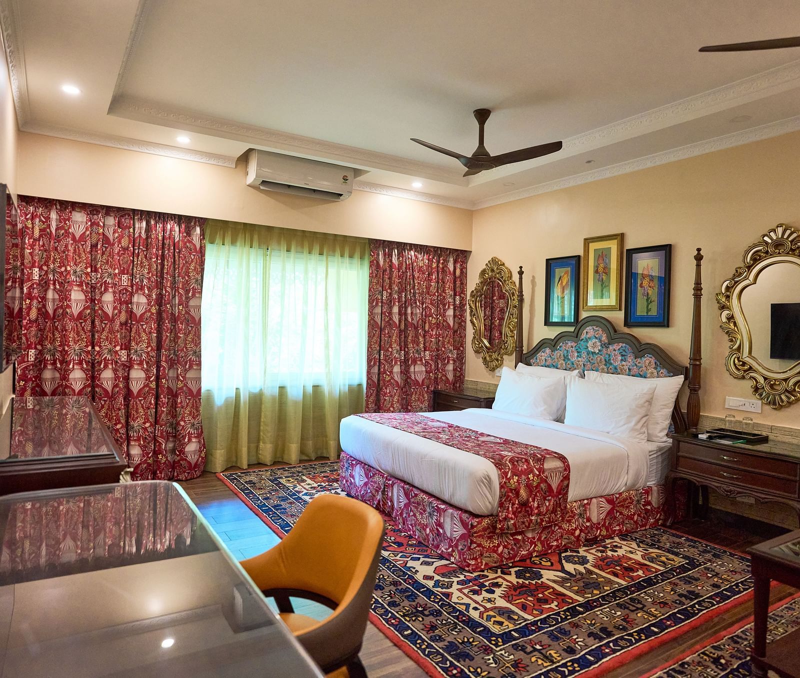 A spacious room featuring a large bed, a desk with a chair, wooden bedside tables, and windows draped with curtains at Mayfair Manor, Alibag