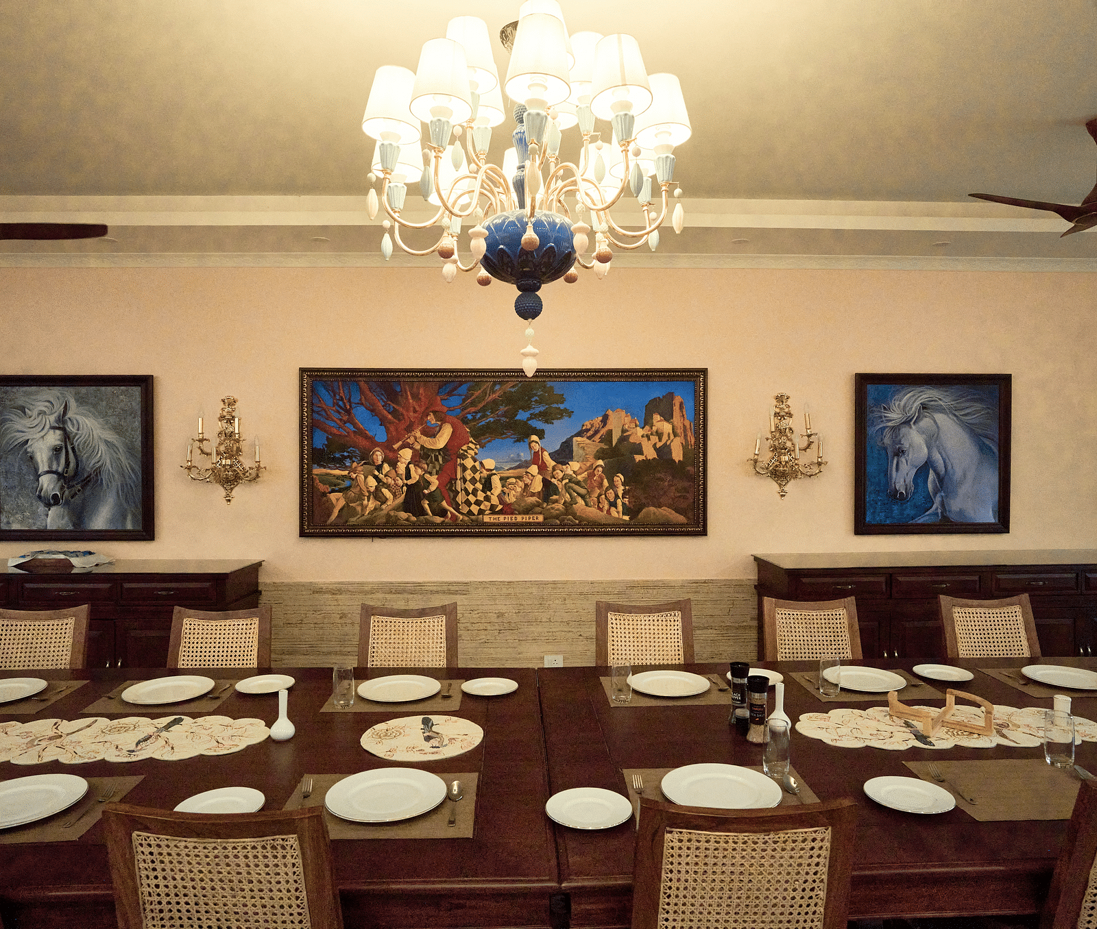 A large dining table elegantly set for a culinary experience, complemented by wall-mounted paintings and a luxurious chandelier overhead at Mayfair Manor, Alibag