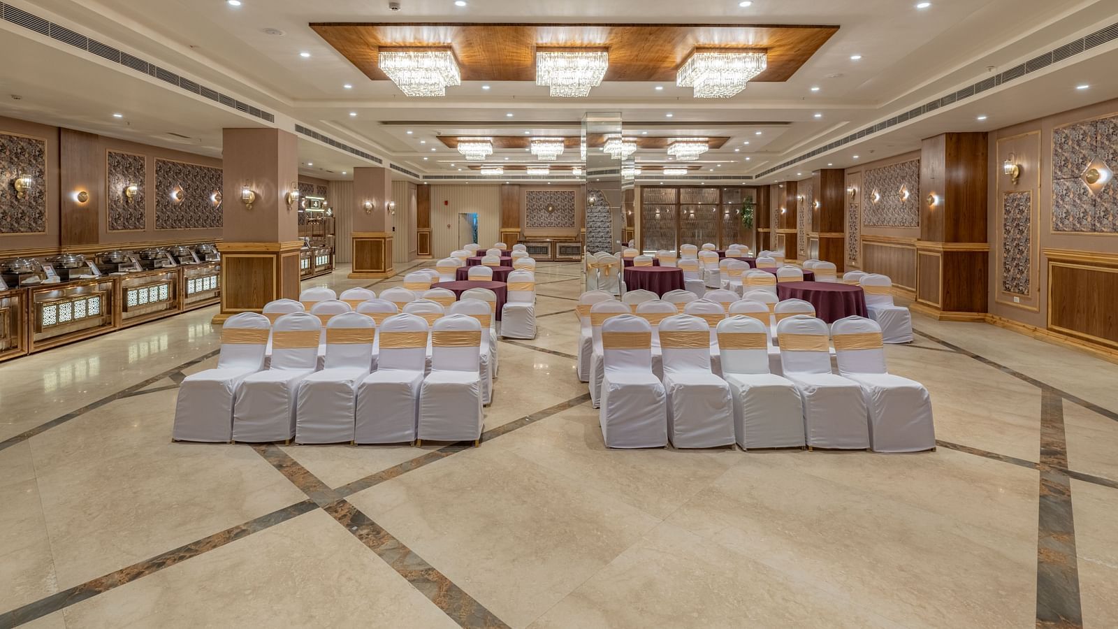 A grand, spacious banquet hall is set up with many round tables covered in white cloths, ready for an event, under a brightly lit, ornate ceiling.