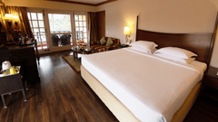 A room with a double bed, wooden furniture & lamps & a large ceiling to floor window with a seating space. Find your ideal Kodaikanal room-booking available now at The Clarton.