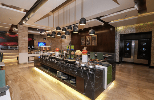 A restaurant bar area at Park Plaza Ludhiana featuring a long counter with pendant lights wooden flooring track lighting on the ceiling and colorful decor in the background.
