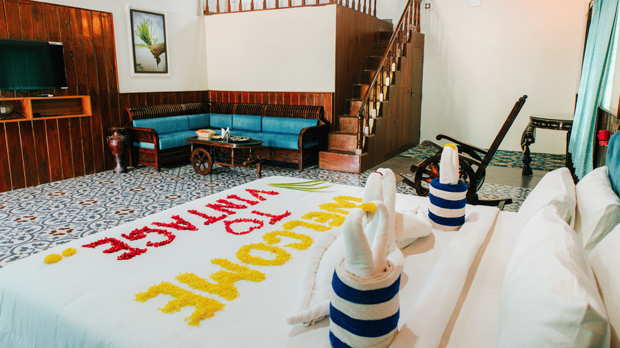 A bedroom view featuring a bed with towel art and text made of petals in the foreground, and a living area with a sofa, TV, and stairs in the background | Vintage Trails By Ilara