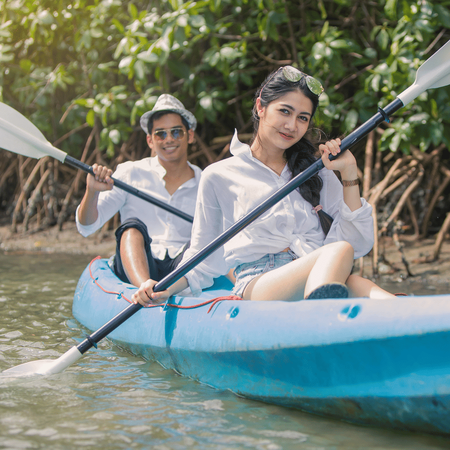 2 individuals, each in a kayak, paddling through calm river waters surrounded by dense mangroves, showing a peaceful and serene exploration setting.