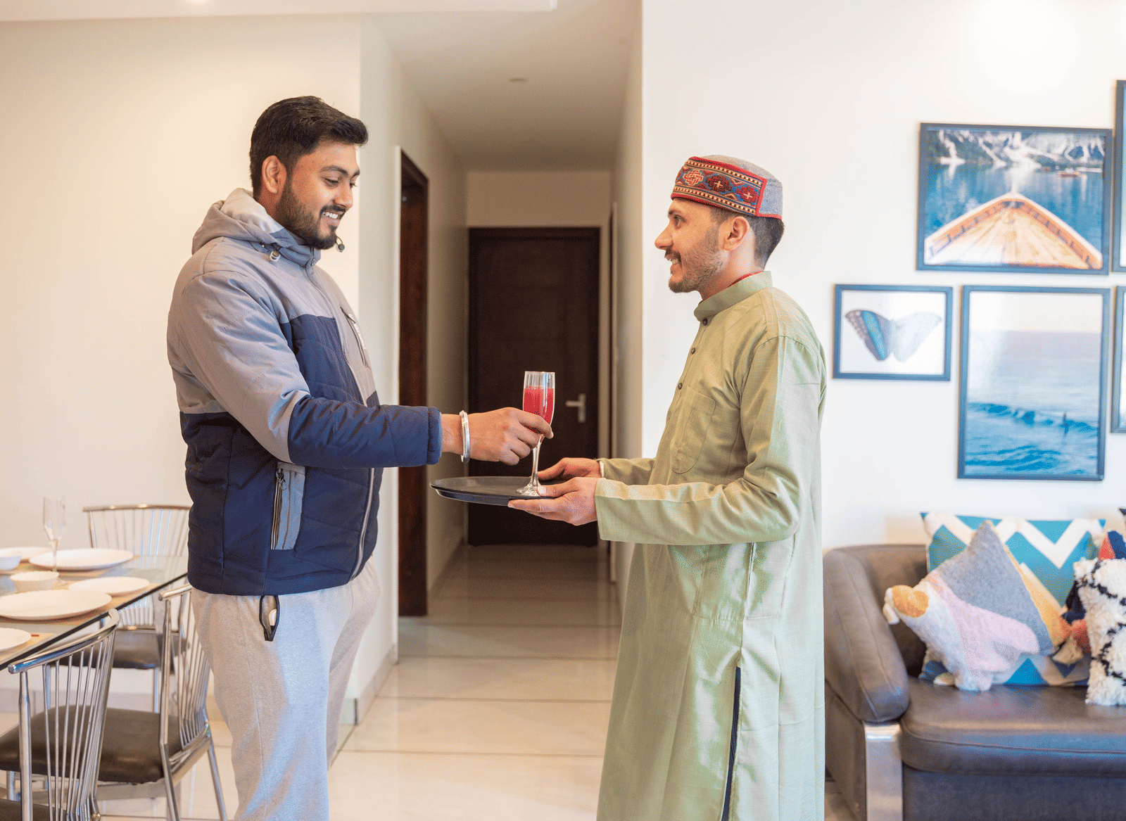 A person receives a beverage from another person inside a room with a dining table, sofa, and wall art at the Perfectstayz Value Shimla (Namah Retreat).