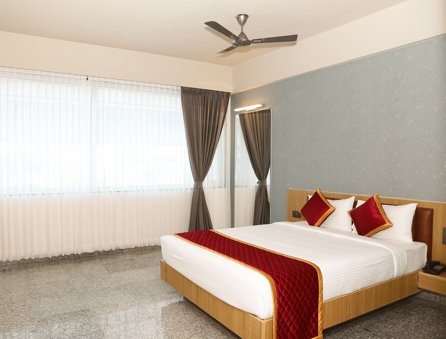 An angled view of a double bed, a large window, and grey curtains at Icon Express By Bhagini, Mahadevapura.