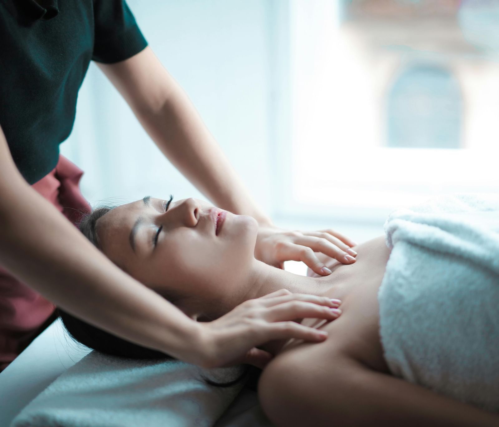 A young woman with her eyes closed is receiving a relaxing neck and shoulder massage from a masseuse.