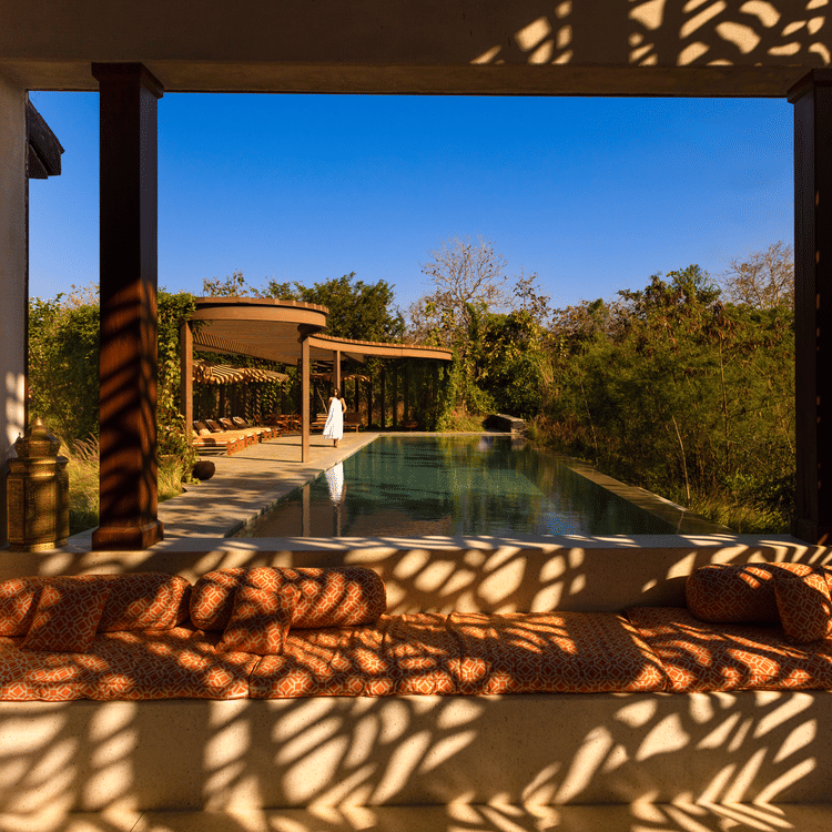 Open lounge area at Aramness, with dappled sunlight creating intricate shadow patterns on the seating and walls, overlooking a private swimming pool surrounded by lush greenery and forest views.