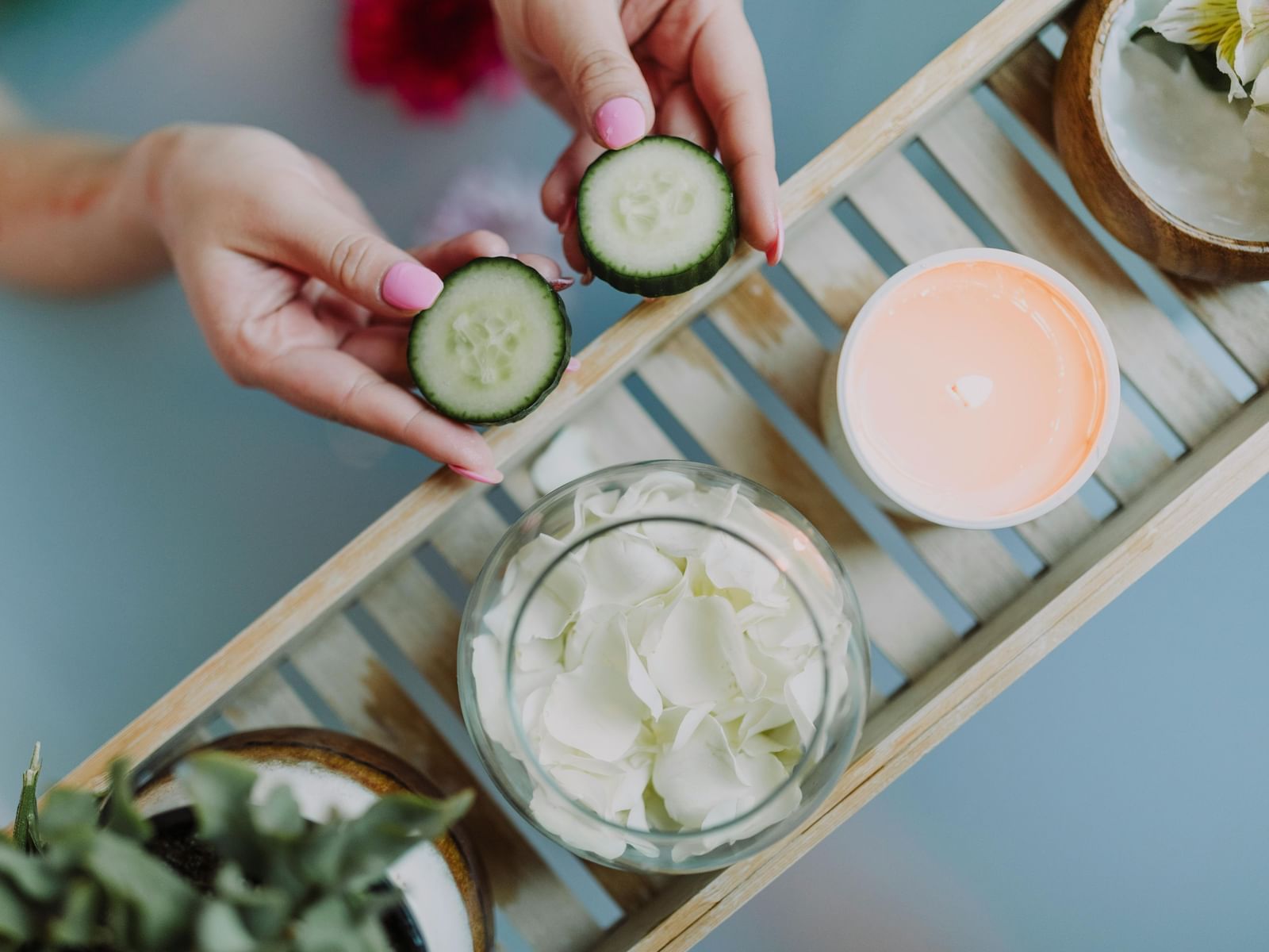 Overhead view of hands showcasing slices of cucumber over a light blue bathtub. A wooden bath candy holds a candle and flower petals.