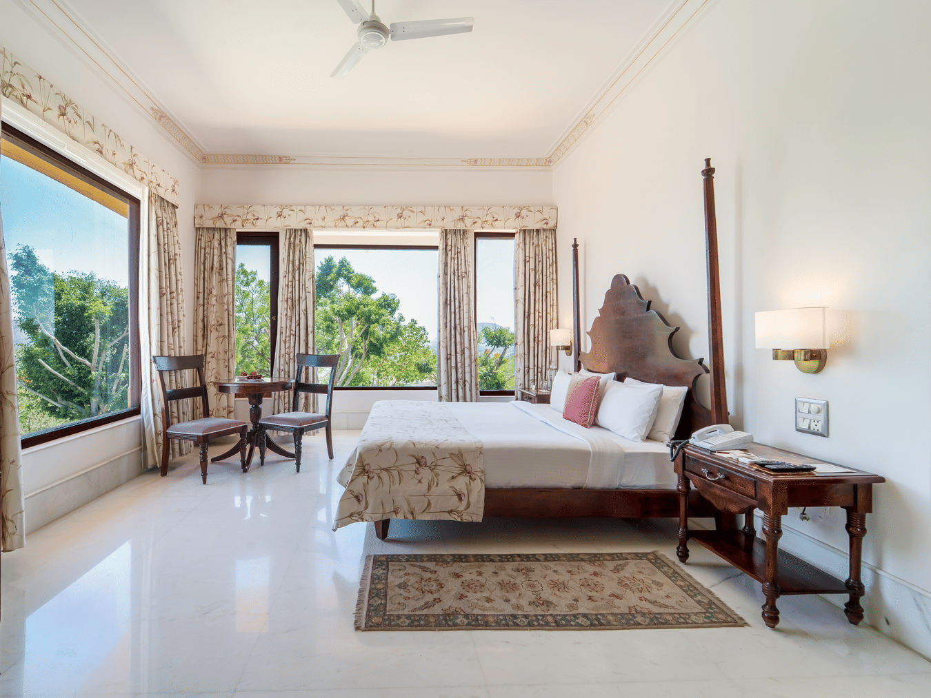 A spacious bedroom featuring a dark wood 2-poster bed, elegant drapery, and large windows with garden views - Fateh Garh, Udaipur