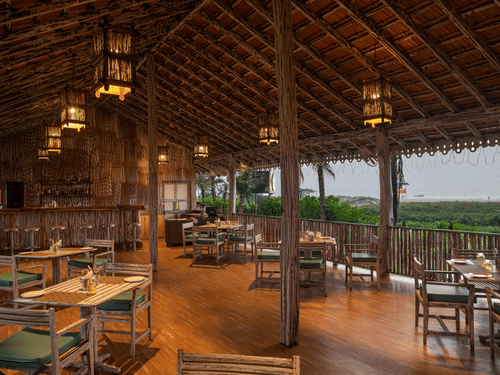 The Kai Seaside Restaurant at Kenilworth Resort & Spa, Goa, featuring a thatched roof, hanging lanterns, wooden tables, and a scenic view of the beach.