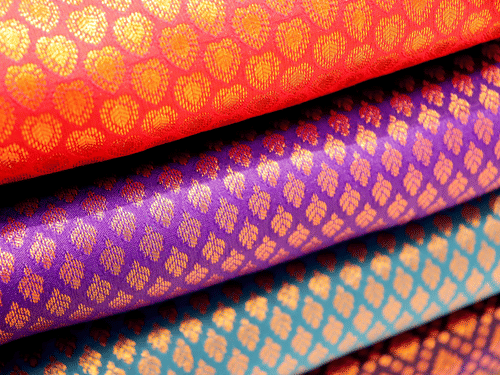 Multiple folded saree pieces with patterned textile designs in different colours stacked on top of each other.