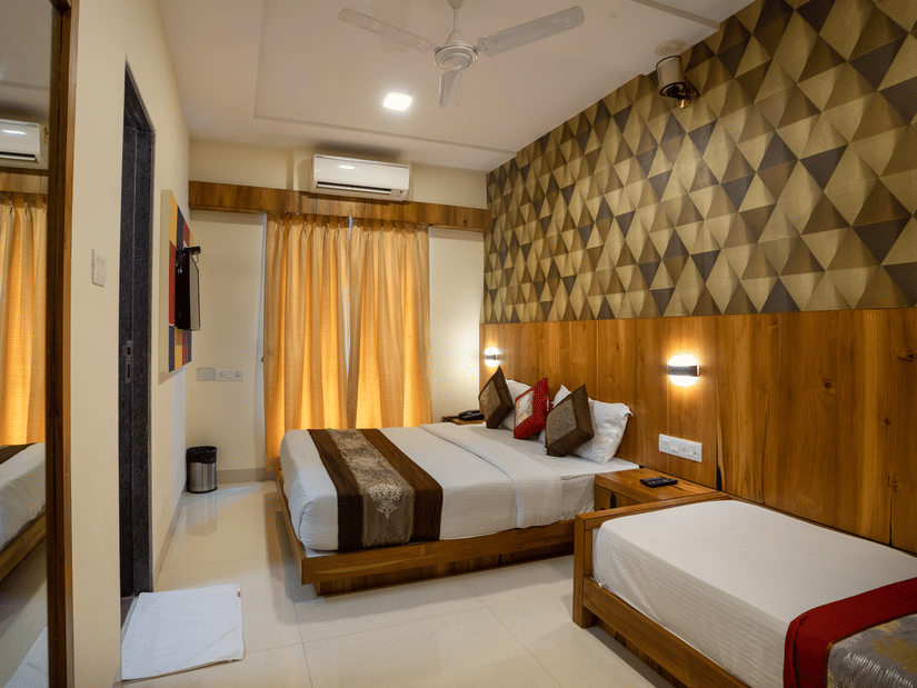 A bedroom at Home2 India with 2 beds, patterned wall panel, wooden accents, bedside lighting, a window with curtains, and a mirror near the entrance.