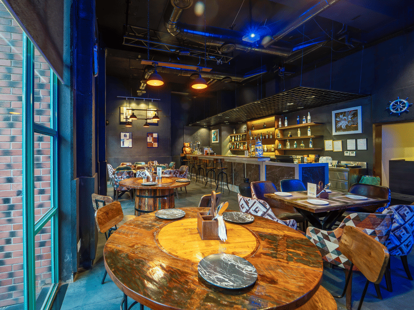  Interior of Rosetum Anjuna's Speakeasy Bar featuring rustic wooden tables, eclectic chairs, a fully stocked back bar, industrial pendant lighting, and blue ambient lighting.