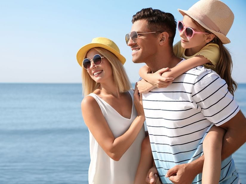A family on a beach, with a woman and a man giving a piggyback ride to a child, all wearing hats and sunglasses, facing the ocean.
