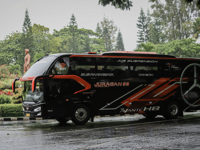 A modern black and orange coach bus driving on a wet road beside a landscaped garden with greenery and trees in the background on a rainy day.