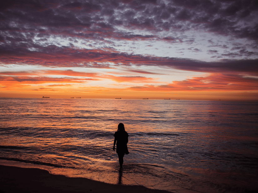 A silhouette of a person standing on the beach against a sunset sky painted in shades of orange, purple, and blue.
