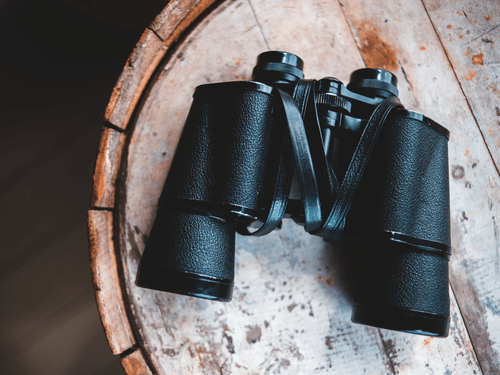 Black binoculars placed on a round wooden surface, with textured tabletop and dark background visible around the object.