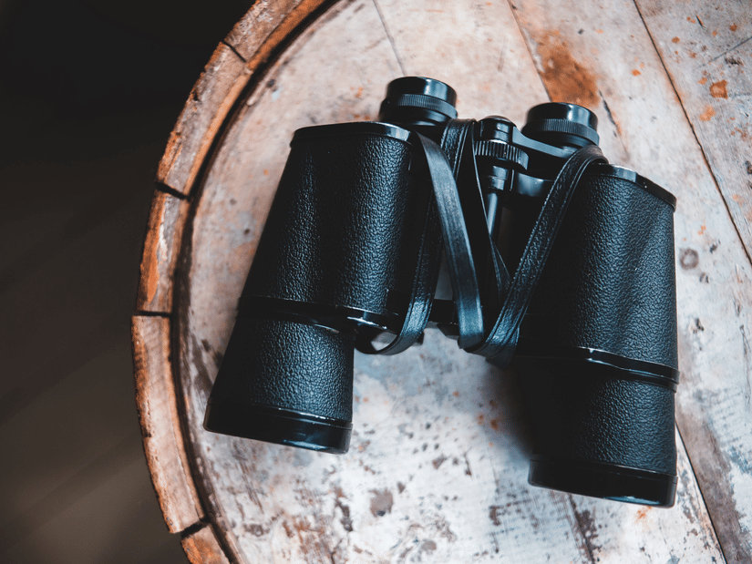 Black binoculars placed on a round wooden surface, with textured tabletop and dark background visible around the object.
