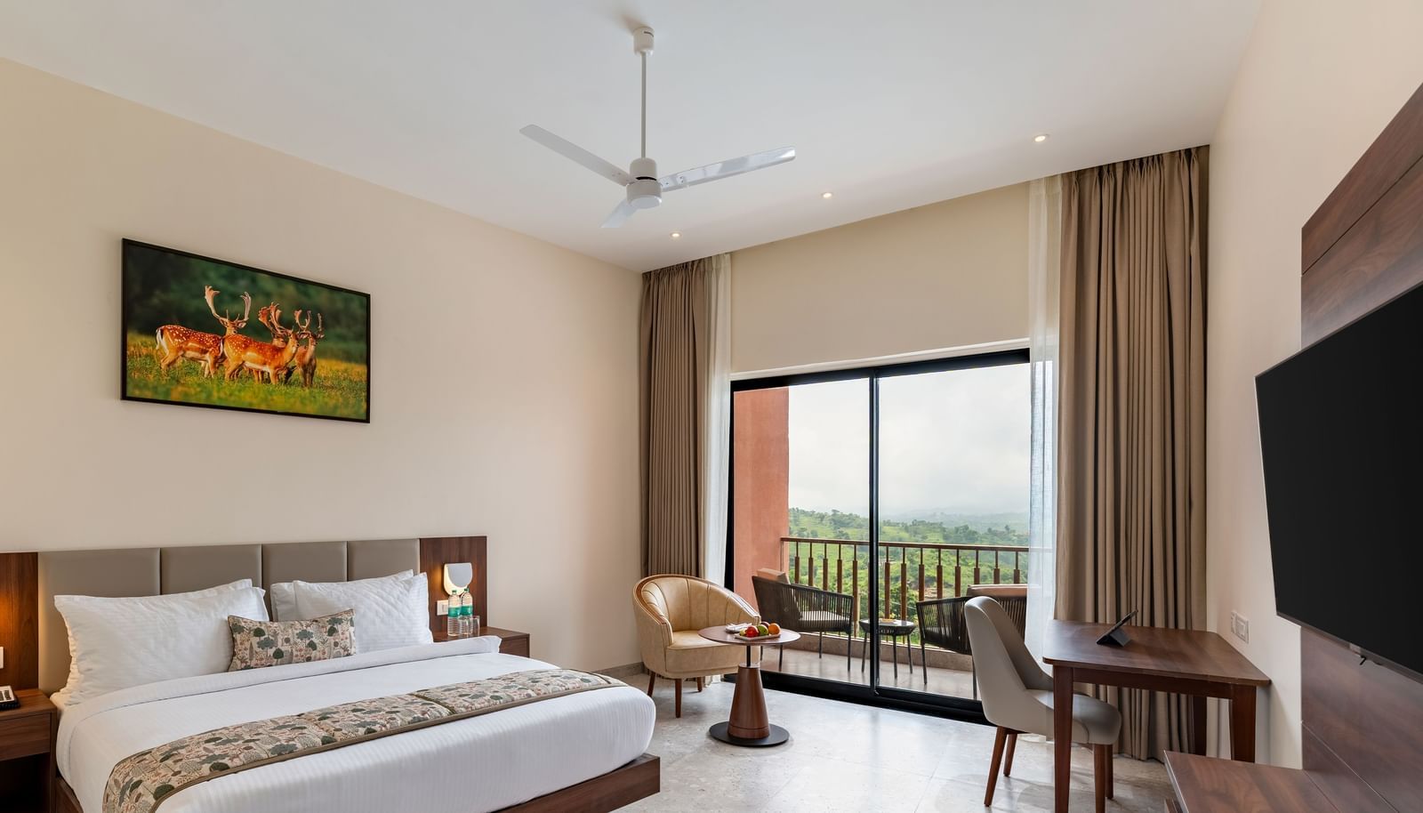 A spacious bedroom at Osian Valley Resort & Spa with a large bed, a desk and chair, a wall-mounted television, and a balcony with a scenic view.