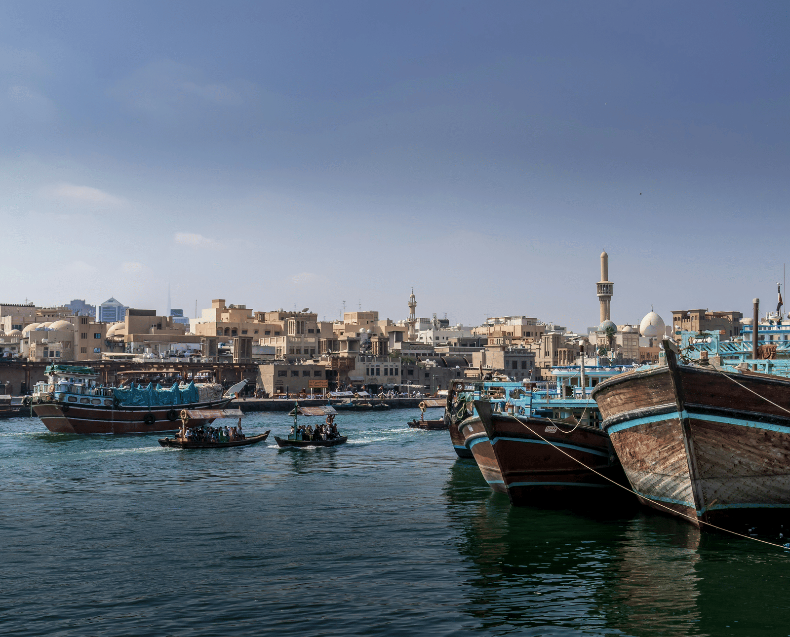 Traditional wooden abra boats docked along a waterfront with city buildings and a mosque visible in the background.
