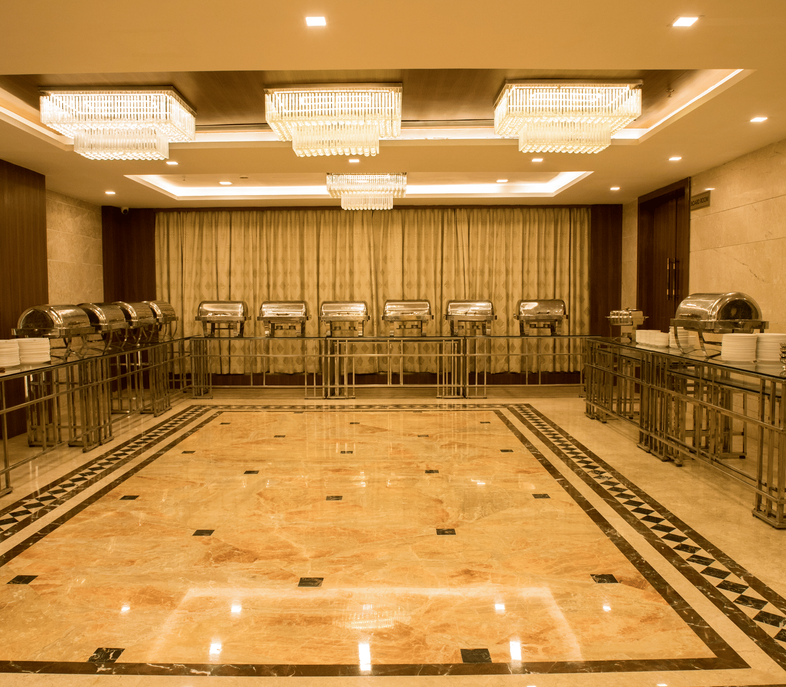 A brightly lit banquet hall with several chafing dishes lined at the far end of the hall at Benzz Park, Vellore.