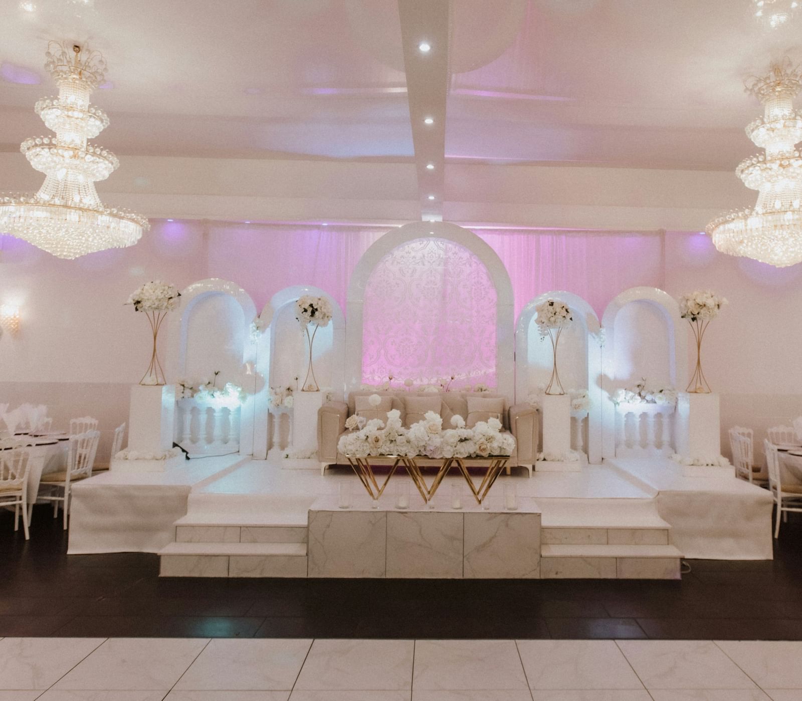 Elegant indoor banquet hall with white chairs, chandeliers, and a floral-decorated stage.