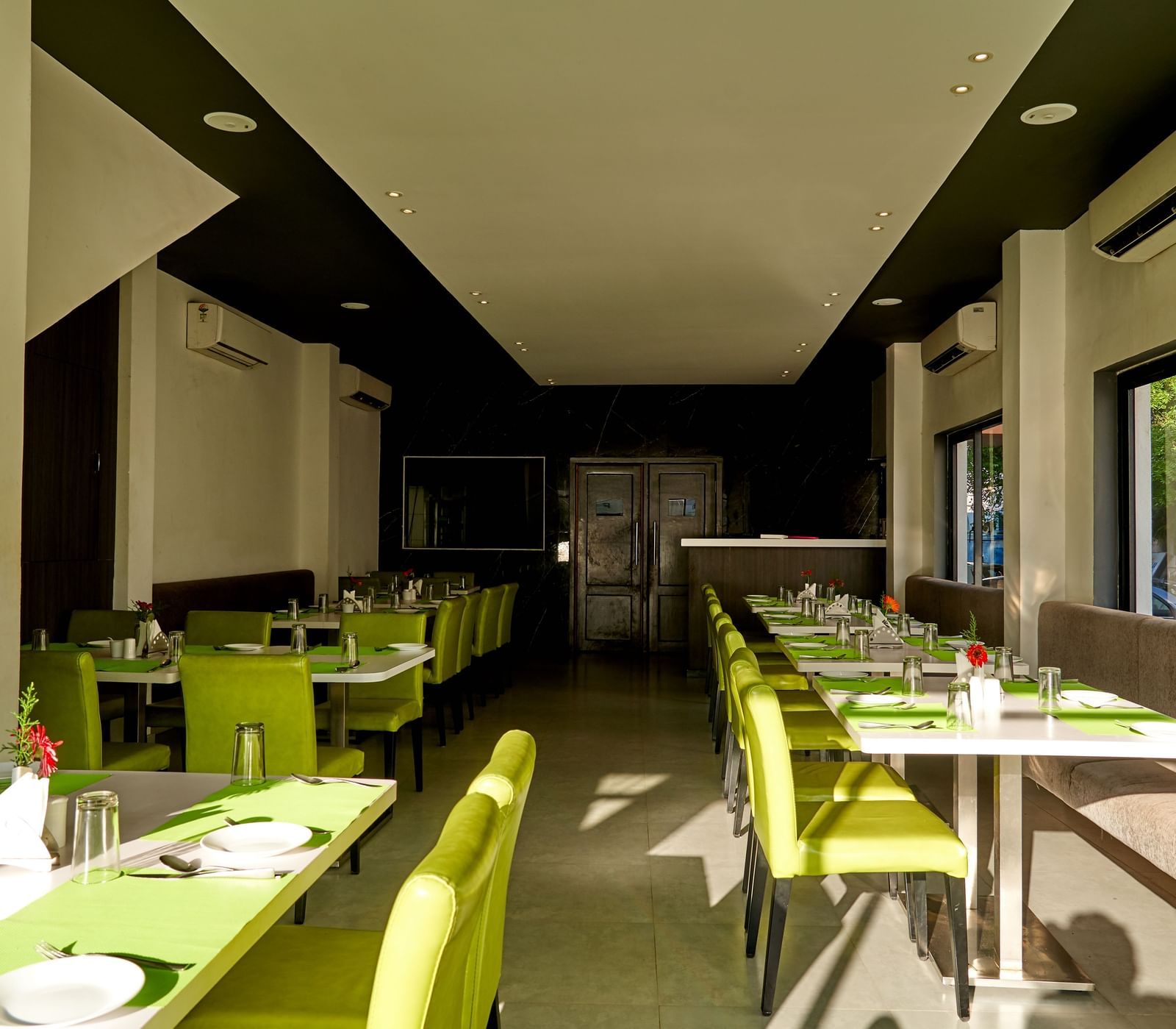 A wide view of a modern dining space at Hotel Mamallaa Heritage, featuring tables with green place settings and a contemporary, bright aesthetic.