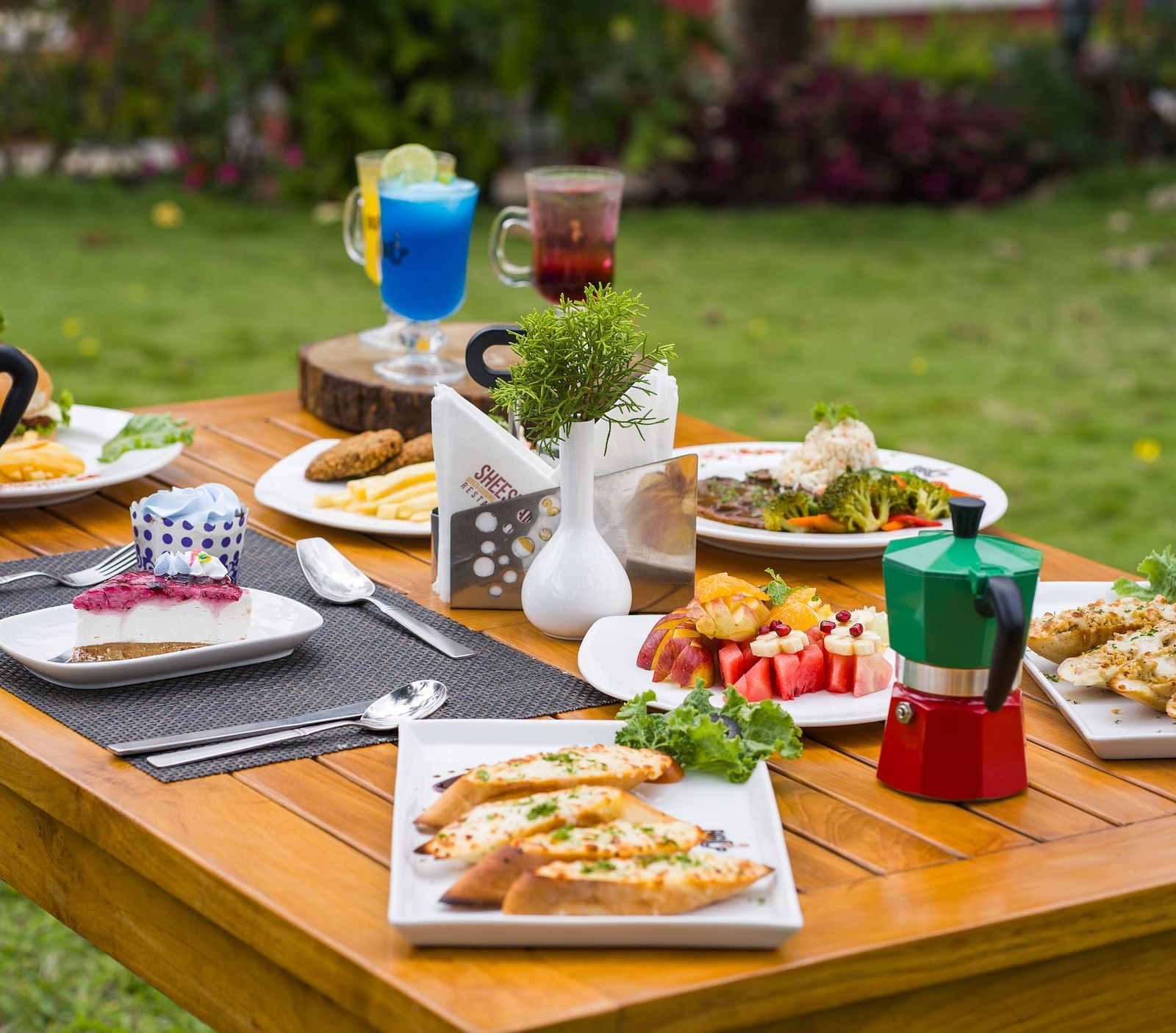 A wooden table is set outdoors with a variety of dishes and drinks, surrounded by a green lawn | Woodstock Resorts, Coorg