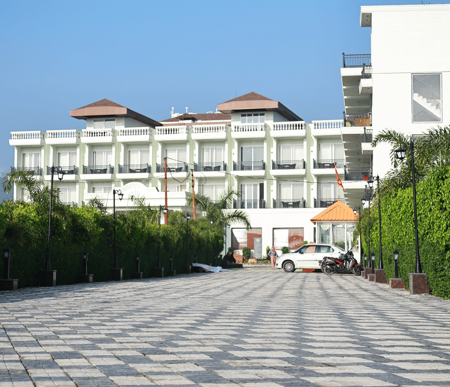 Long paved walkway lined with green hedges leading toward the main white hotel building and parking area.
