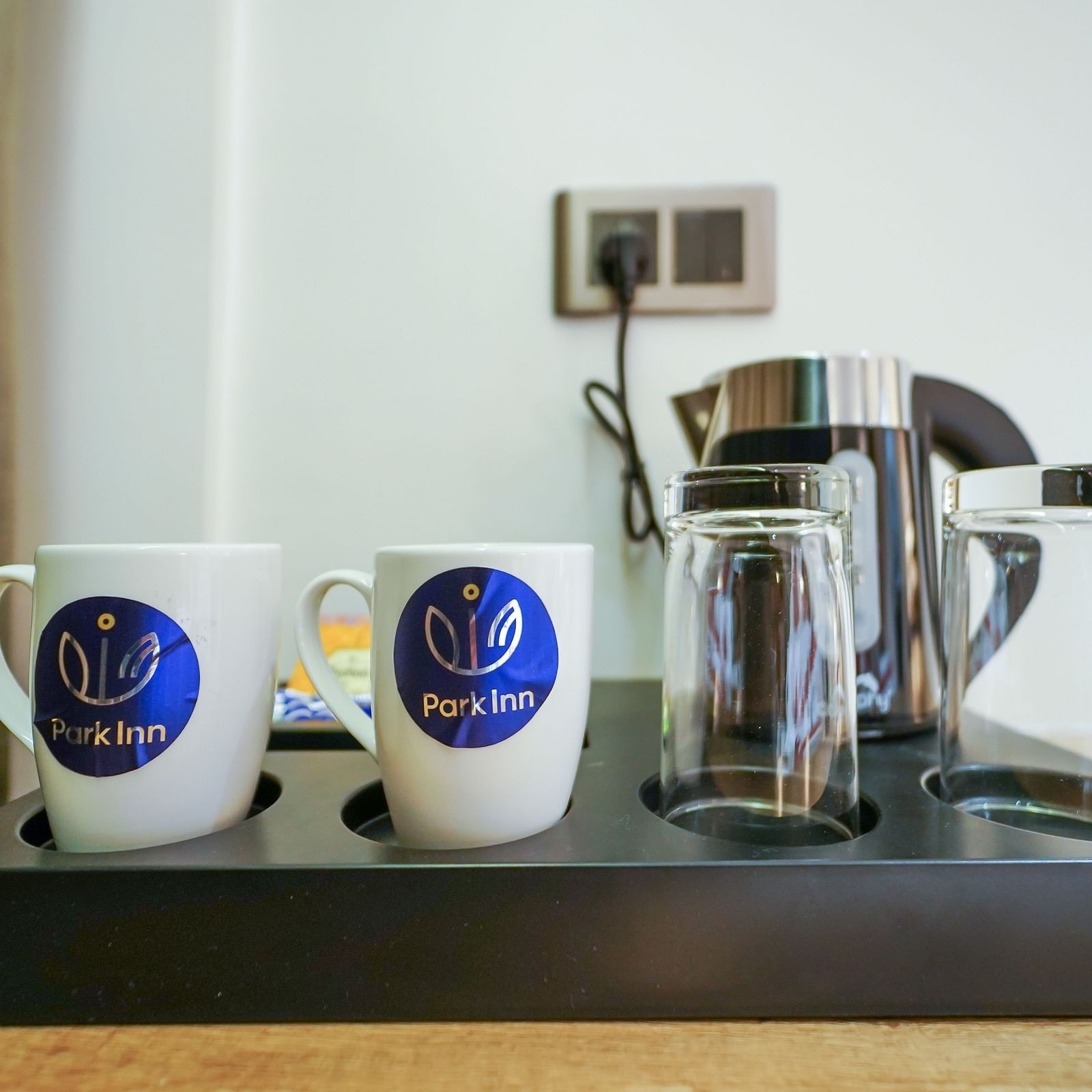 A hospitality tray holds two mugs with logos, a glass kettle, and two drinking glasses on a light-toned surface near a wall socket - Hotel Park Inn, Nellore.