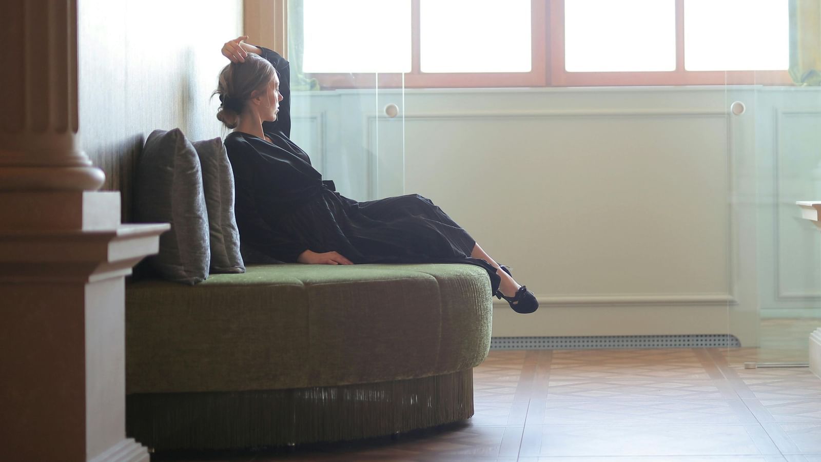 Person sitting on a cushioned bench in a hallway with tall pillars and soft lighting, gazing out towards a large window.