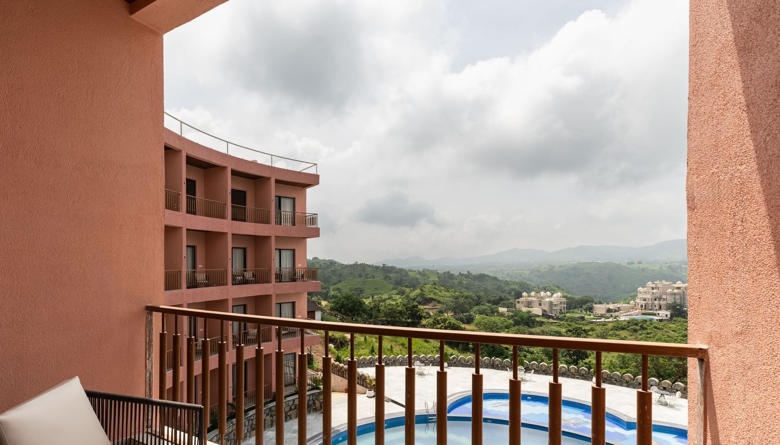 A balcony at Osian Valley Resort & Spa, furnished with 2 chairs, overlooking a swimming pool and a valley with green hills.