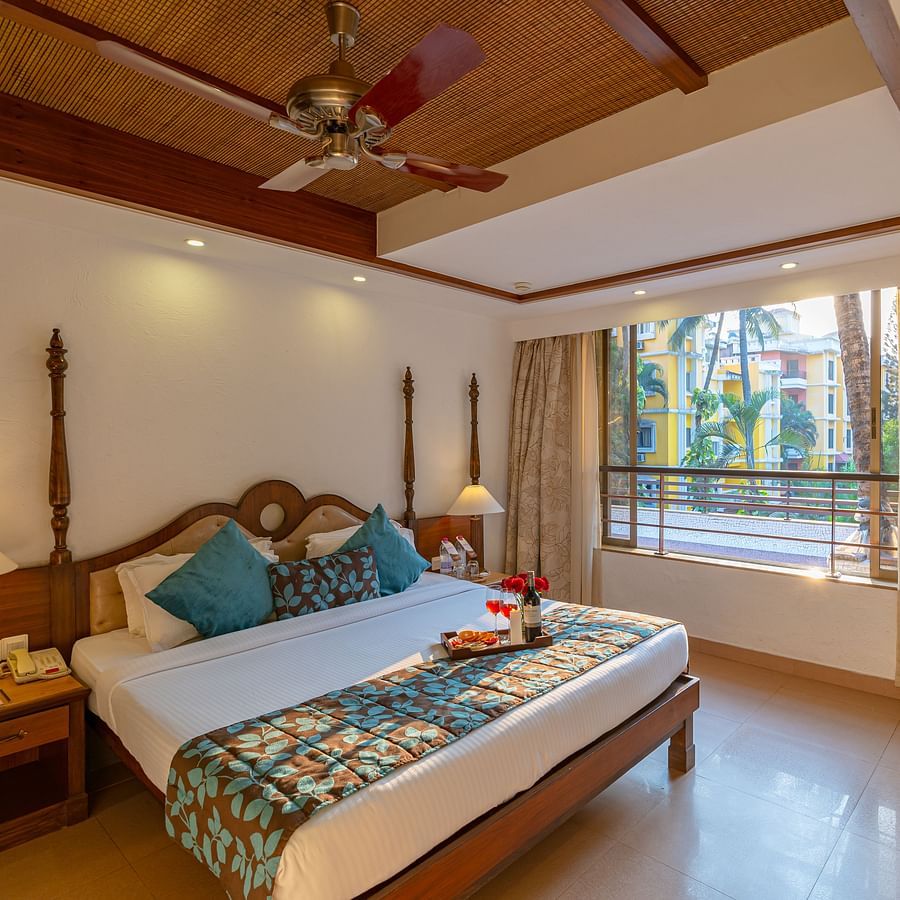 An image of a room featuring a neatly made bed, a fan, two table lamps, side drawers, and a large window.