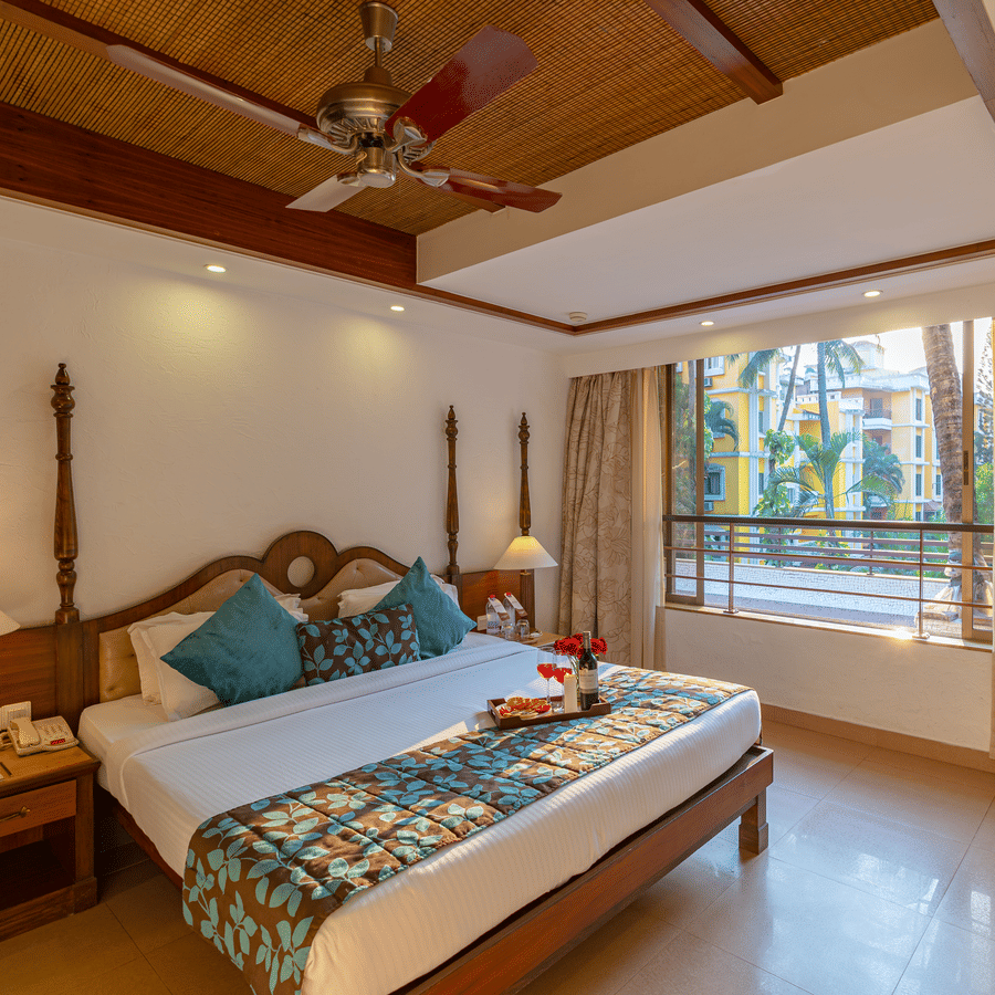 An image of a room featuring a neatly made bed, a fan, two table lamps, side drawers, and a large window.