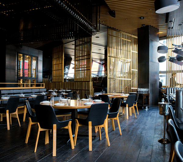 An interior view of a modern restaurant with dark flooring, wooden tables, and dark blue chairs arranged neatly.