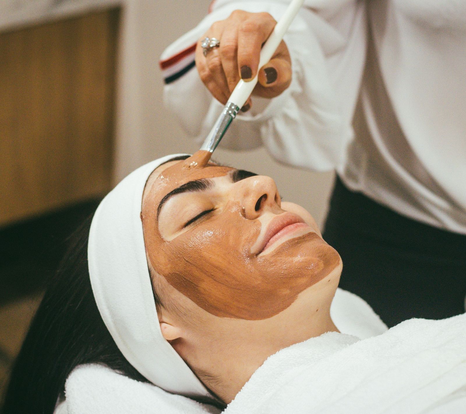 A woman lying down with her eyes closed enjoying a mud mask being applied on her face with the help of a brush.