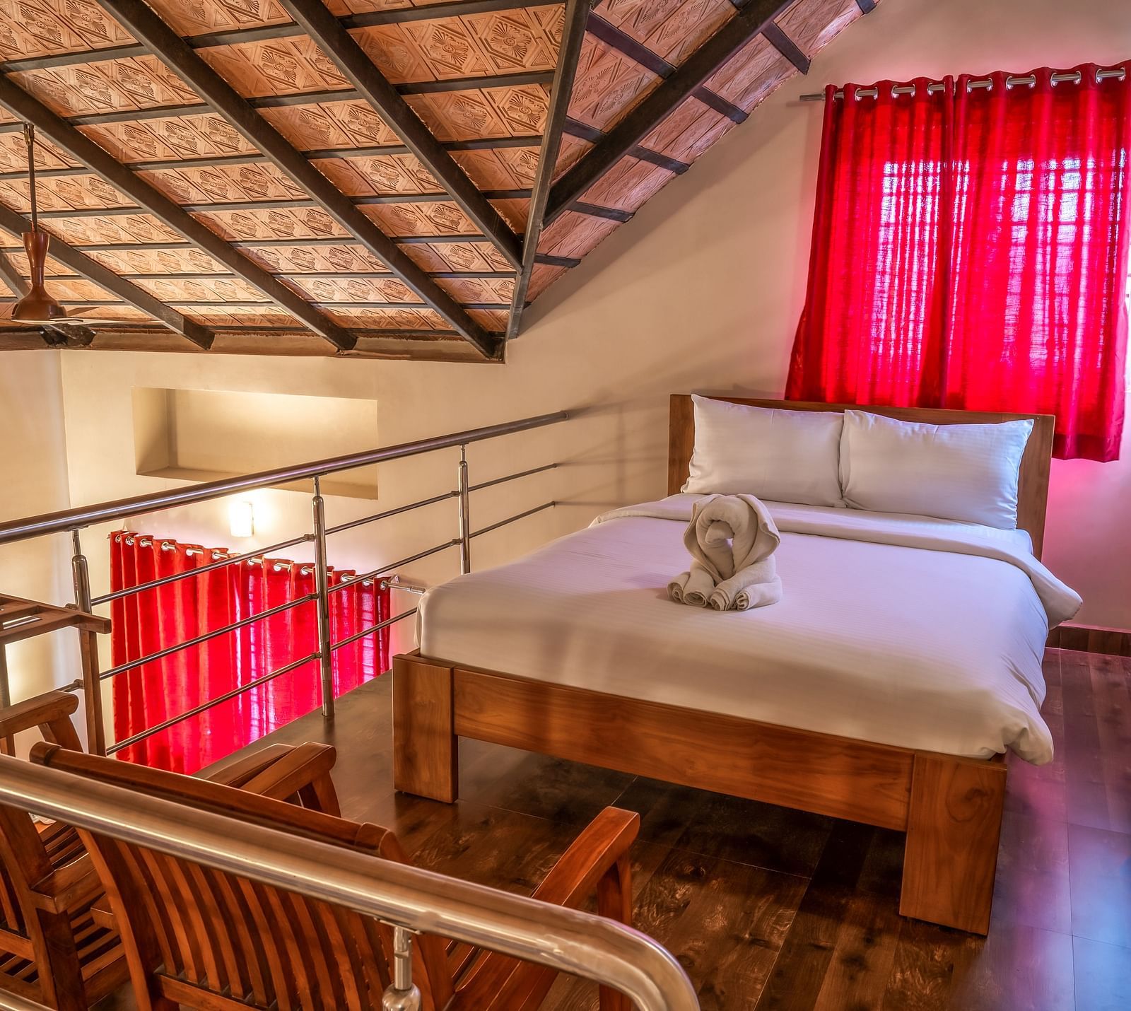 An elevated view shows Attic Villa featuring a wooden bed frame, white linens, and red curtains, with a glimpse of the room's upper level | Woodstock Resorts, Coorg