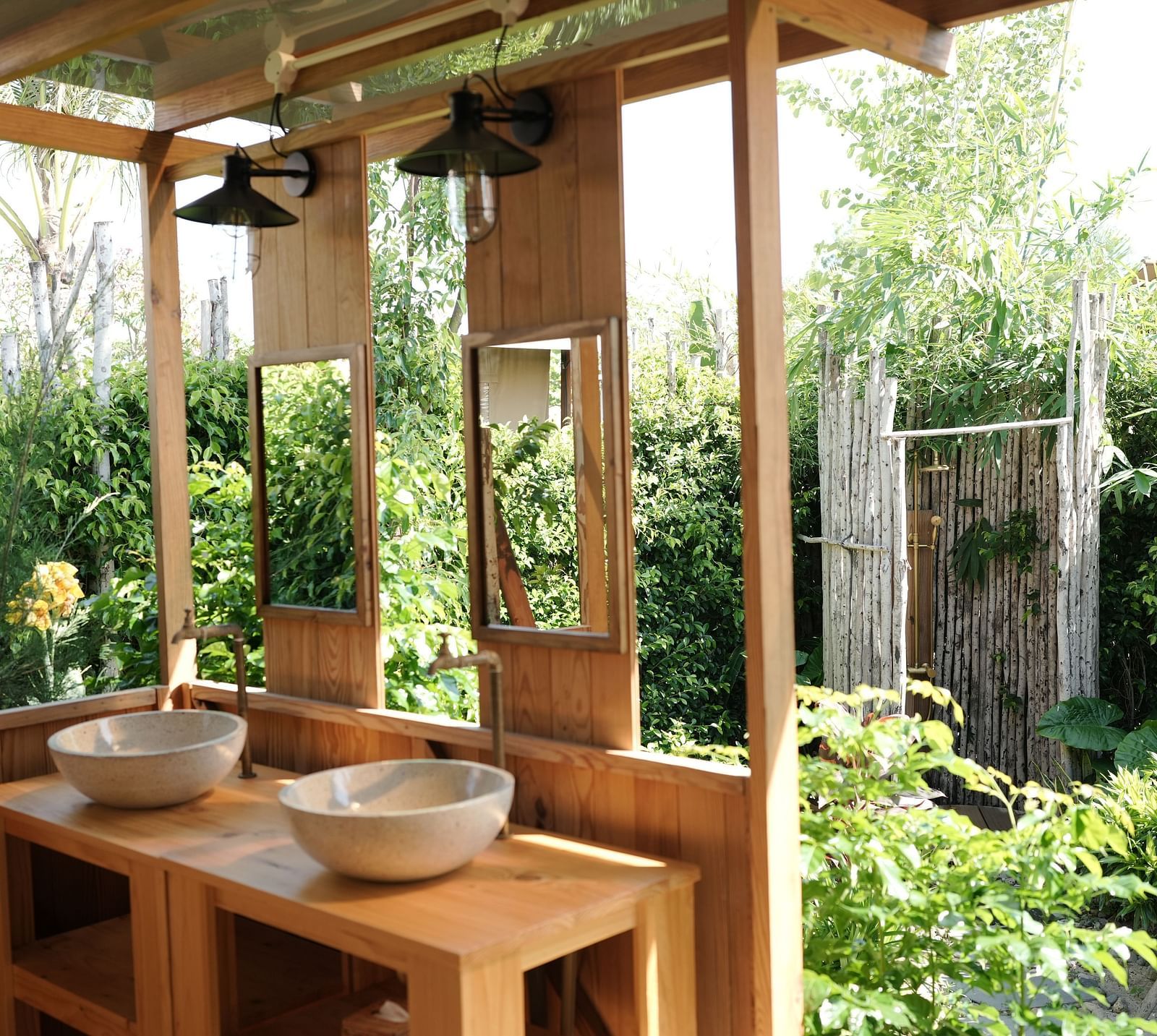  An outdoor bathroom area at WoCAL Retreat Ke Ga, part of the Mindful Tent, featuring twin wooden washbasins, mirrors, and surrounded by lush greenery.